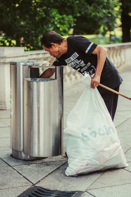 In the foreground of the image, a large pile of mixed waste and rubbish is overflowing from a metal communal bin designated for paper and cardboard disposal, situated on a paved area. The trash includes various items such as crumpled cardboard boxes, plastic bags, paper packaging, and other miscellaneous waste materials, some spilling onto the surrounding pavement. To the right of the bin, there are additional black and red rubbish bags, with some material partially exposed, alongside flattened cardboard boxes. Behind the bins, a blue truck or van is visible, indicating possible private waste collection services. In the background, there is a commercial building with a blue metal scaffold, glass windows, and a sign for a fish bar, with storefronts visible at ground level, suggesting an urban or suburban environment. The overall scene depicts an on-site rubbish accumulation, reflecting the importance of regular waste removal services such as those provided by Waste Disposal Maida Vale for maintaining cleanliness and preventing clutter accumulation.