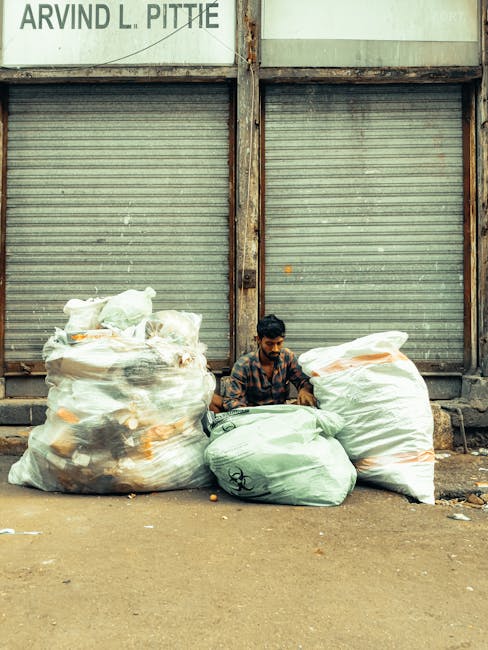A man with dark hair and a beard is seated on the ground in front of a row of large, partially transparent plastic rubbish bags filled with mixed waste, including visible paper, plastic, and organic materials. The bags are white with orange and black markings and are positioned against a weathered, corrugated metal shutter door that is closed. The background features a metal frame structure with signs of rust, supporting the shutters and a faded sign with partially visible text at the top left. The scene is set outdoors on a paved surface with some scattered debris, suggesting a waste collection or disposal area. The lighting appears natural, with even daylight illuminating the scene. The presence of waste bags and the setting relate to rubbish removal activities, possibly indicating the need for professional waste services like those from Waste Disposal Maida Vale, and shows a typical private or independent approach to waste handling outside formal rubbish collection points.