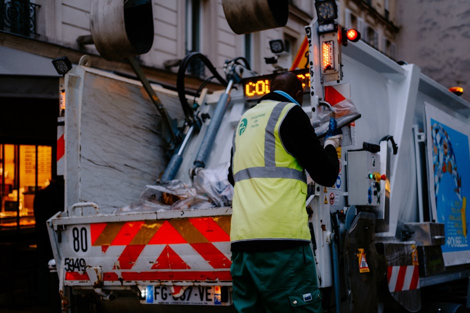 A waste management worker wearing a high-visibility yellow vest with reflective stripes and dark trousers is operating a large municipal rubbish collection vehicle during low-light conditions, with an urban residential building visible in the background. The worker is seen from behind, using controls on the side of the vehicle, which is painted white with red and orange hazard markings. The vehicle's rear section is filled with a mixture of waste materials, including crumpled paper, plastic, and other refuse, with some of it piled up near the open container. The scene captures elements of an independent or commercial rubbish collection process, typical of private waste disposal services such as Waste Disposal Maida Vale, highlighting on-site clearance rather than traditional curbside collection. The environment appears urban, with warm ambient lighting from nearby windows and streetlights, emphasizing the operational context of professional waste removal services in Maida Vale area.