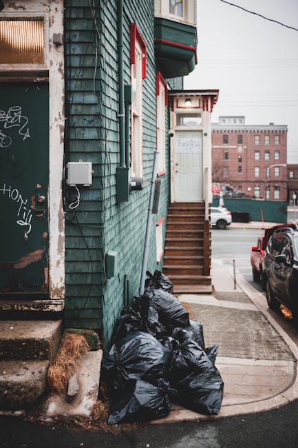 A cluster of black plastic rubbish bags is piled against the exterior wall of a teal-colored building with wooden shingle cladding, situated on a residential street. The bags are tightly packed and appear to be filled with household waste, sitting on a small concrete pathway adjacent to a set of wooden steps leading up to a white door with a decorative overhang. Next to the bags, a weathered metal utility box is mounted on the wall, with a white cable running from it. The lower portion of the building shows signs of wear, including chipped paint and some graffiti on a nearby door frame. In the background, parked cars and the facades of other brick buildings are visible, indicating an urban environment typical of local rubbish collection areas. The scene is illuminated by natural daylight, highlighting the textures of the building’s shingles and the shiny plastic surface of the rubbish bags, illustrating a typical private waste removal scenario managed by services such as Waste Disposal Maida Vale as an alternative to council collection.