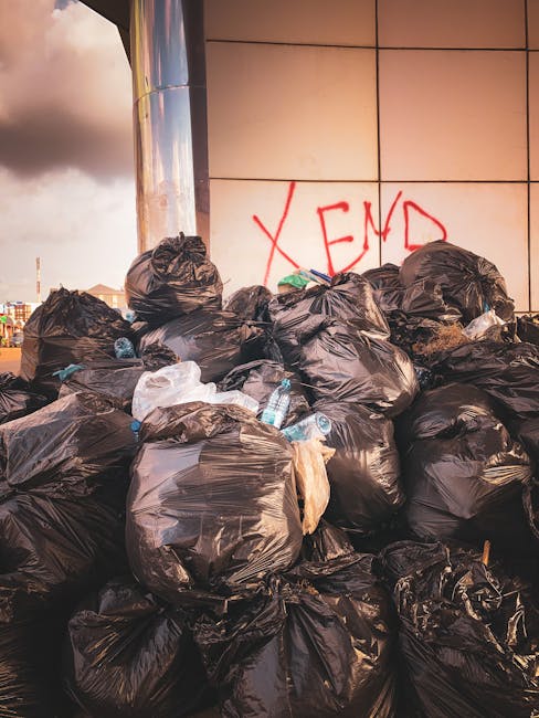 A large pile of black trash bags filled with waste sits against a modern building with a reflective metallic surface and beige tiled wall. Some of the bags are partially torn or bulging, revealing glimpses of waste inside. Among the bags, there are several clear plastic bottles and scattered debris, indicating mixed refuse. Behind the pile, graffiti spelling 'XEND' is painted in red on the tiled wall, suggesting an area where private or alternative waste disposal occurs outside typical municipal collection. The scene is outdoors, with stormy clouds in the background, and the lighting suggests late afternoon or early evening. The setting conveys an environment where independent rubbish removal, such as services offered by Waste Disposal Maida Vale, may be needed to manage excess waste generated in urban areas. The contrast between the reflective building surface and the dark, bulky refuse emphasizes the urban nature of the rubbish accumulation, highlighting the importance of professional waste removal solutions for maintaining cleanliness and order in busy neighborhoods.