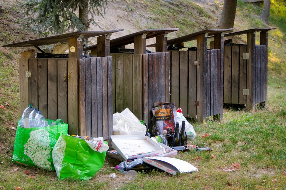 A cluster of black plastic rubbish bags is piled against the exterior wall of a teal-colored building with wooden shingle cladding, situated on a residential street. The bags are tightly packed and appear to be filled with household waste, sitting on a small concrete pathway adjacent to a set of wooden steps leading up to a white door with a decorative overhang. Next to the bags, a weathered metal utility box is mounted on the wall, with a white cable running from it. The lower portion of the building shows signs of wear, including chipped paint and some graffiti on a nearby door frame. In the background, parked cars and the facades of other brick buildings are visible, indicating an urban environment typical of local rubbish collection areas. The scene is illuminated by natural daylight, highlighting the textures of the building’s shingles and the shiny plastic surface of the rubbish bags, illustrating a typical private waste removal scenario managed by services such as Waste Disposal Maida Vale as an alternative to council collection.