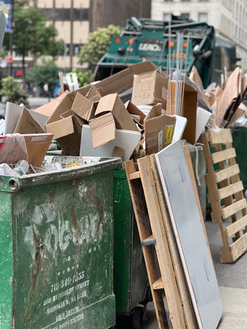 A middle-aged man with dark hair, wearing a black T-shirt with white printed text on the front, is seen leaning forward to dispose of rubbish into a stainless steel public waste bin located on a paved sidewalk. The bin features a shiny metallic surface, with multiple compartments for different types of waste, and is positioned near a low concrete or stone railing. Behind the man, lush green trees and foliage are visible, indicating an outdoor urban setting during daylight hours. He holds a large white plastic bag, which appears to be filled with various rubbish, and uses a wooden stick or broom handle to help guide the bag into the bin. The scene highlights the act of private rubbish disposal, as part of an independent rubbish collection or on-site waste clearance effort facilitated by Waste Disposal Maida Vale, with the environment maintained tidy and clean through active waste management practices.