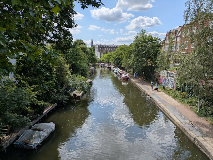 A narrow urban canal lined with tall, leafy trees on both sides, with reflections of the blue sky and scattered clouds visible on the water surface. Parked cars are situated along the right bank, adjacent to a paved footpath where a few pedestrians are walking. On the left side, small boats are moored alongside the water, with some covered by protective tarps. In the background, there are multi-story brick residential buildings with large windows, and a church spire rising above the treetops, indicating a typical cityscape. The overall scene is bright and clear, suggesting a sunny day with natural daylight illuminating the lush greenery, nearby buildings, and the tranquil waterway, which is a common feature in parts of Maida Vale and Little Venice. This image reflects a quiet residential area that might require occasioned rubbish removal services to maintain cleanliness alongside alternative waste handling options, such as private collection by Waste Disposal Maida Vale.