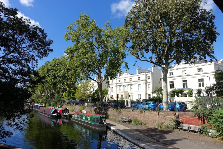 A wide view of a narrow canal lined with large, leafy green trees, with three narrowboats moored along the water's edge, reflecting in the calm surface. On the opposite side of the canal, there is a paved walkway with a few empty wooden benches, and behind the benches, a brick retaining wall supports a row of modern white residential buildings with multiple windows, some with small balconies and potted plants. Several parked cars, including a blue sedan, are visible in front of the buildings, with some greenery and shrubbery along the street. The sky above is bright blue with a few scattered clouds, indicating clear weather during daylight. The scene captures a typical urban riverside environment where private boat mooring and residential property coexist, illustrating the setting where local waste collection services like Waste Disposal Maida Vale might manage rubbish generated in this area.