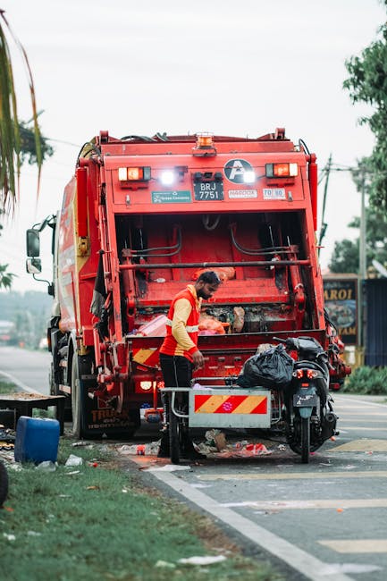 A waste collection worker wearing a high-visibility orange and yellow jacket is operating a large red rubbish disposal truck positioned on the side of a street. The truck's rear loading section is open, revealing mechanical components and storage areas, with some debris and waste material visible nearby on the ground. The worker appears focused, handling waste or engaging in a collection task. Adjacent to the truck, there is a black garbage bag placed on a metal platform, and a small platform or base with a railing supports the worker. In the background, out-of-focus trees, utility poles, and distant signage suggest a suburban or urban environment. The scene is illuminated by natural daylight, with overcast sky and some greenery visible along the roadside, illustrating a typical scene of waste collection or rubbish removal service, possibly part of private waste handling activities supported by Waste Disposal Maida Vale.