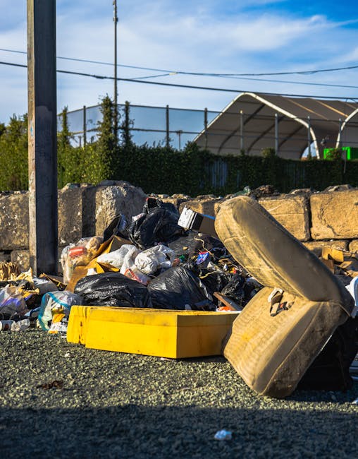A pile of mixed household rubbish and waste materials sits on dark, loose gravel near a stone wall, including black plastic garbage bags, a yellow plastic tub, and an old, dirty beige car seat with fabric upholstery. The waste appears to be recently discarded and partially spilled onto the ground, with some items leaning against the stone wall. In the background, there is a fence, hedge, and a tennis court with a canopy structure, under a partly cloudy blue sky. The scene suggests a private rubbish collection or on-site waste clearance, with Waste Disposal Maida Vale possibly involved in managing such waste removal. The environment appears outdoor, with natural lighting highlighting the clutter, and the setting indicating an informal disposal area or roadside rubbish accumulation, relevant to non-authority based waste handling services.