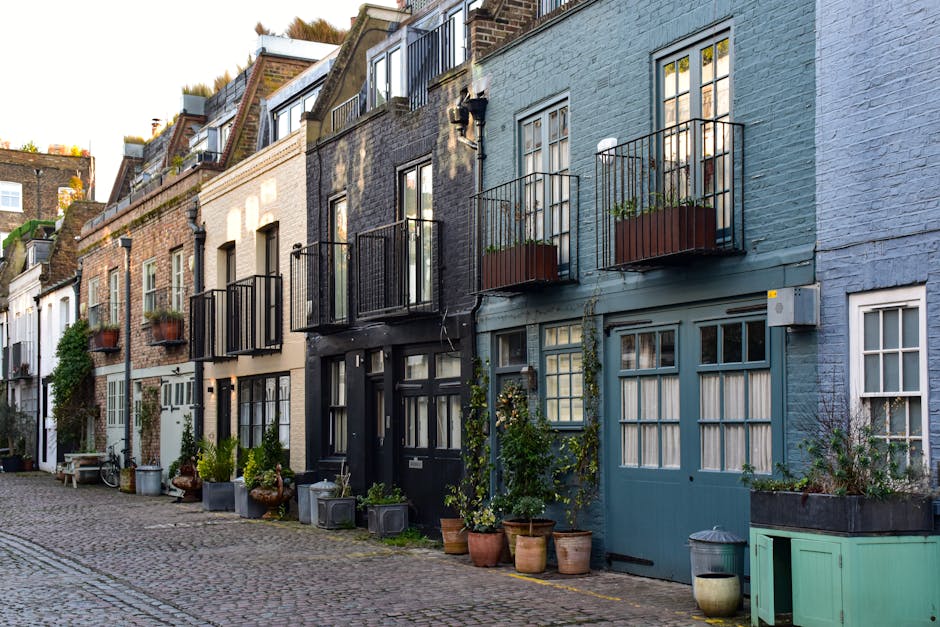 A row of narrow, multi-story residential houses with varying facades along a cobbled street, each featuring small front gardens or potted plants positioned directly outside their entrances. The buildings display a mix of materials, including brick and painted brick finishes; some have textured surfaces while others are smoother. Several houses have black or grey front doors, while others have doors painted in pastel shades, complemented by large windows with multiple panes. Small balconies with black iron railings extend from upper-floor windows, some with potted plants on the railings. The front gardens are decorated with a variety of potted greenery, flowering plants, and garden ornaments, creating a quaint and inviting atmosphere. The street is quiet and appears in natural daylight, with a soft, warm light indicating late afternoon or early evening. The scene reflects an urban area with a community-oriented aesthetic, naturally aligning with services such as waste disposal or rubbish removal that support maintaining cleanliness and orderliness in residential neighborhoods, possibly managed by companies like Waste Disposal Maida Vale.
