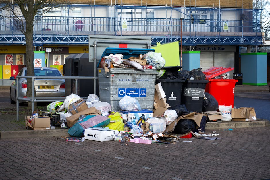 A large collection of mixed household waste and recyclable materials, including cardboard boxes, plastic bags, paper, and discarded packaging, piled beside a series of wheelie bins and rubbish containers in an urban outdoor setting. The waste is spilling onto a paved area adjacent to a metal barrier, with some items stacked on top of each other and others scattered randomly. The bins include a grey bin labeled for mixed paper and cardboard, a black bin, and a bright red bin, all positioned on a concrete curb. Behind the waste, there is a multi-storey building with blue safety barriers and scaffolding, and storefronts with signage, indicating a commercial or retail area. The scene suggests an instance of excess rubbish accumulation that might require professional waste removal services, such as those provided by Waste Disposal Maida Vale, focusing on efficient and responsible rubbish clearance in the Maida Vale area.
