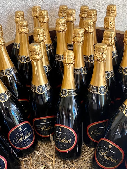 A group of champagne bottles arranged upright on a bed of straw or wood shavings, with black glass bodies and gold foil wrapping around the necks and tops. The bottles feature labels with a dark background and gold lettering, displaying the brand name 'Ardau' and the logo, which appears to be a gold crest or emblem. The bottles are closely packed together, filling the frame from foreground to background, creating a dense cluster. The background consists of a plain, light-colored wall, possibly textured. The lighting is even, highlighting the shiny, reflective surfaces of the bottles and the crinkled texture of the gold foil. This image might relate to private event supplies or celebratory packaging, fitting within contexts of waste disposal or collection of packaging materials, as seen in waste management or rubbish removal services offered by Waste Disposal Maida Vale. The scene emphasizes the presentation of packaged products likely destined for disposal or recycling in a waste management context.