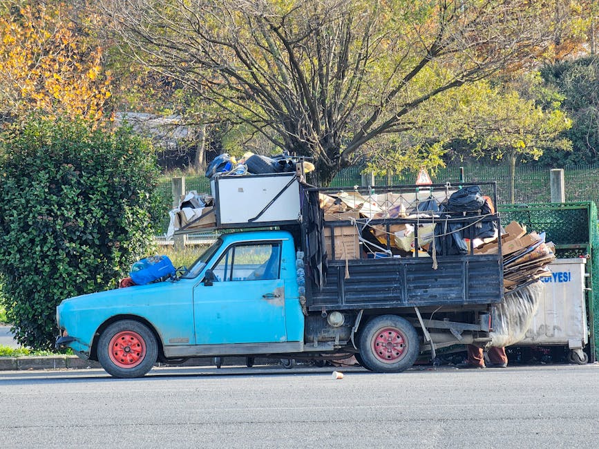 A small, old blue utility truck parked on the side of a street, loaded with various types of rubbish and waste materials. The truck's black cargo area is filled with flattened cardboard boxes, black trash bags, and miscellaneous debris, some of which are loosely secured with ropes. The vehicle's wheels are painted red, and the front of the truck shows minor signs of wear, with visible rust and chipped paint on the bodywork. Behind the truck, a large, leafy tree with a mix of green and yellow foliage extends over a fence, indicating an outdoor environment possibly in a residential or urban area. The scene is illuminated by natural daylight, with shadows cast on the pavement. The truck appears to be in the process of an independent waste collection, possibly arranged through a private waste disposal service such as Waste Disposal Maida Vale, supporting alternative rubbish removal options outside local authority collections.