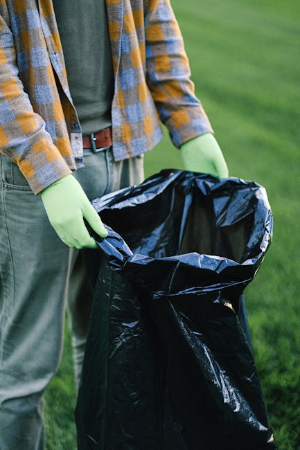 A person wearing a checkered shirt in shades of yellow, blue, and grey, along with green rubber gloves, is holding open a large black plastic rubbish bag outdoors. The bag appears to be empty or nearly so, with its opening wide to accommodate waste. The person stands on a paved or dirt surface, with a background of a well-maintained grassy area. The scene is lit by natural daylight, suggesting a clear day. The surrounding environment hints at an outdoor setting suitable for waste collection or rubbish removal. The focus is on the individual's torso and hands, with no face visible. This image illustrates aspects of refuse collection, possibly linked to a private or independent rubbish disposal service, such as those offered by Waste Disposal Maida Vale, and emphasizes the importance of proper handling and disposal of waste materials in a domestic or outdoor setting.
