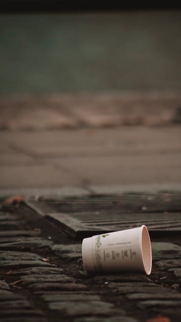 A large green wheeled rubbish bin filled with red and black bags of waste, placed outdoors on a paved surface next to a dark brick wall. The bin has a white label with black and green text, and is positioned on a wooden pallet. The bags contain flexible door seals, as indicated by visible text on the packaging. The scene suggests an onsite waste collection setup, possibly for commercial or large-scale domestic rubbish disposal, with additional bins visible nearby. The environment appears to be a back alley or side yard area, typical of private property waste management arrangements. The lighting is natural, illuminating the textured surface of the brick wall and the materials within the bin. Waste Disposal Maida Vale may handle such refuse collection and disposal tasks, including private waste handling or alternative rubbish removal methods, consistent with the context of rubbish management services.