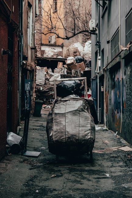A collection of overflowing rubbish bins and scattered waste materials situated on a paved sidewalk in front of a commercial building. The scene includes a large grey mixed waste bin with a blue recycling label for paper and cardboard, which is overflowing with crumpled papers, cardboard boxes, and assorted packaging. To the right, there are additional black and red waste bins, some of which are also filled to capacity with black plastic bags and rubbish spilling over. Surrounding the bins, various discarded items such as paper, plastic containers, cardboard boxes, and packaging are scattered across the ground, contributing to an untidy appearance. The background features a building with retail storefronts, including signage and windows, and construction scaffolding on the upper part of the building, indicating ongoing external maintenance or renovation. A grey metal railing runs parallel to the waste area, and a parked vehicle is partially visible behind the bins. The overall scene highlights a typical urban rubbish accumulation, suitable for a discussion on private waste collection or rubbish removal services such as those provided by Waste Disposal Maida Vale.